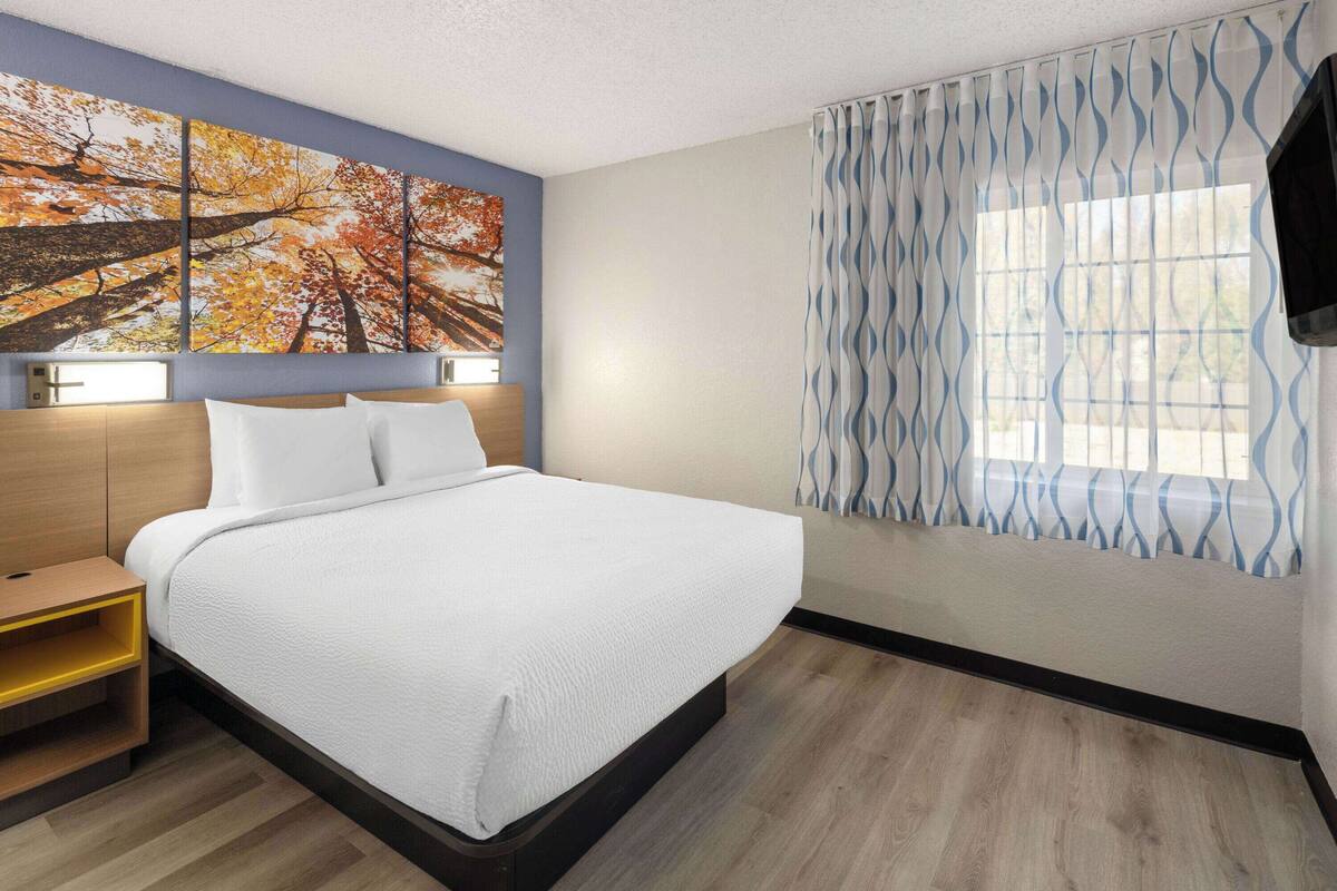 A hotel room with a bed, a nightstand, a wall-mounted TV, and a window with patterned curtains.
