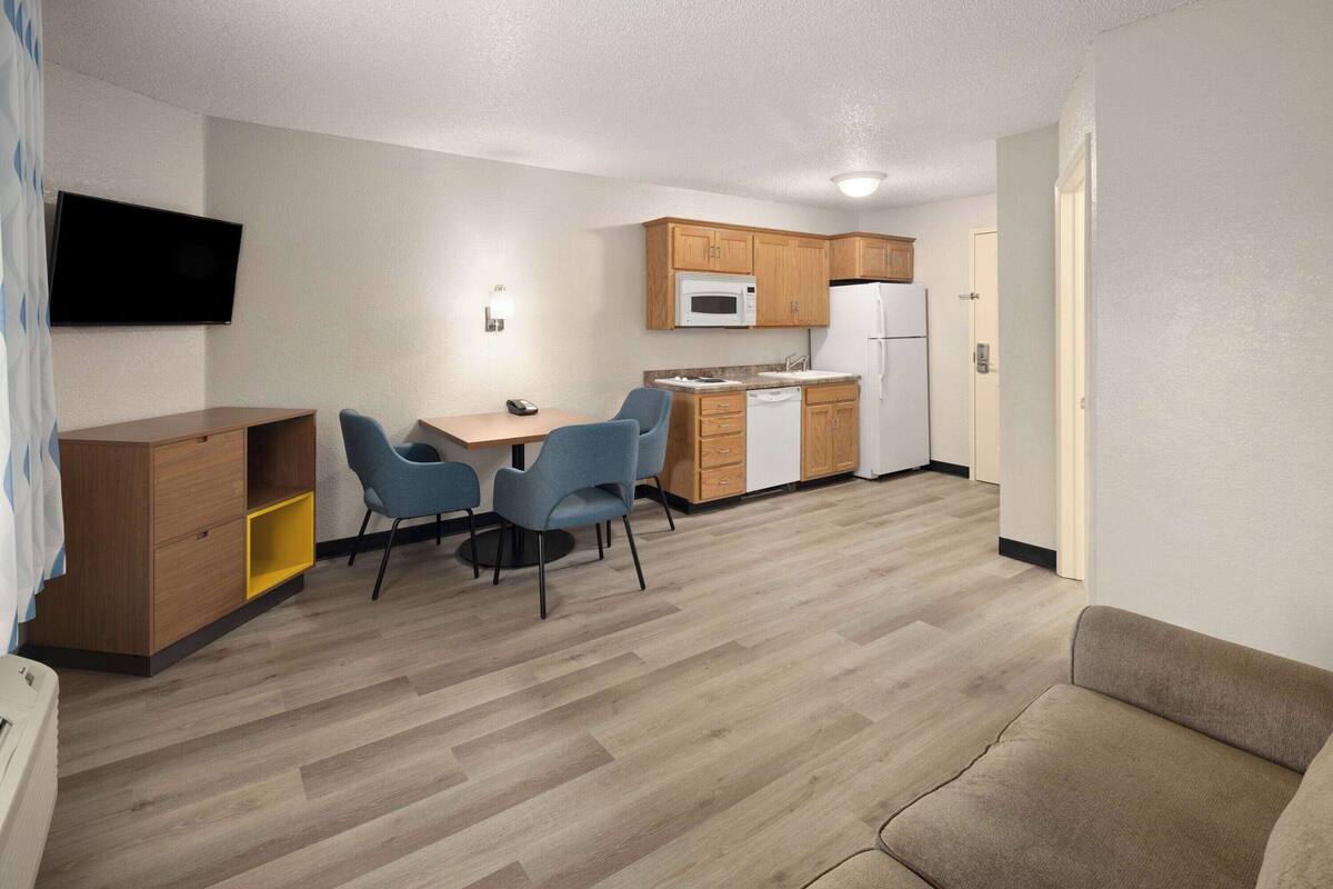 A compact living space with a kitchenette, a dining area with blue chairs, a sofa, and a flat-screen TV mounted on the wall.