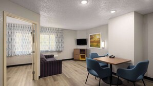 A modern hotel room with a wooden floor, a sofa, a dining table with chairs, a TV mounted on the wall, and a painting. - Days Inn & Suites by Wyndham Green Bay WI. (Green Bay)