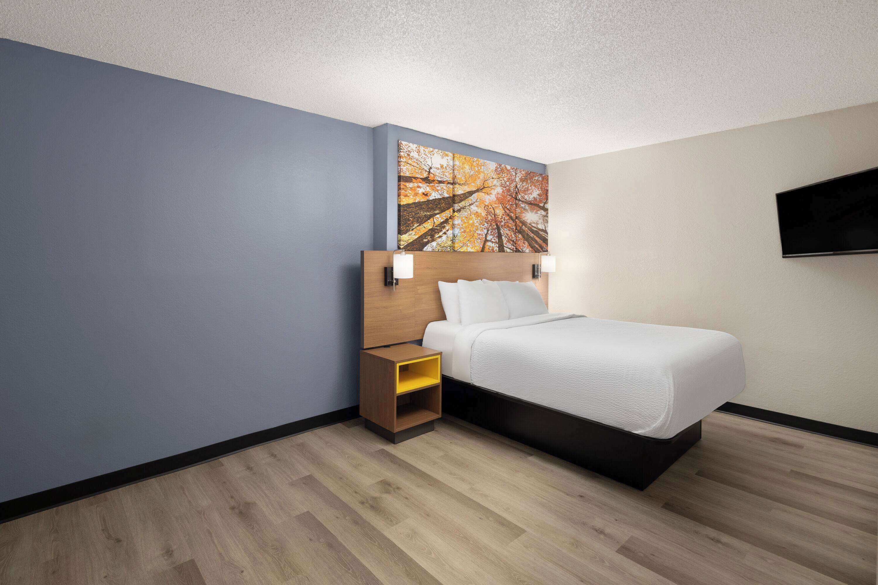 Una habitación de hotel moderna con cabecera de madera, una cama individual, una mesita de noche con estante amarillo y un televisor de pantalla plana montado en la pared.