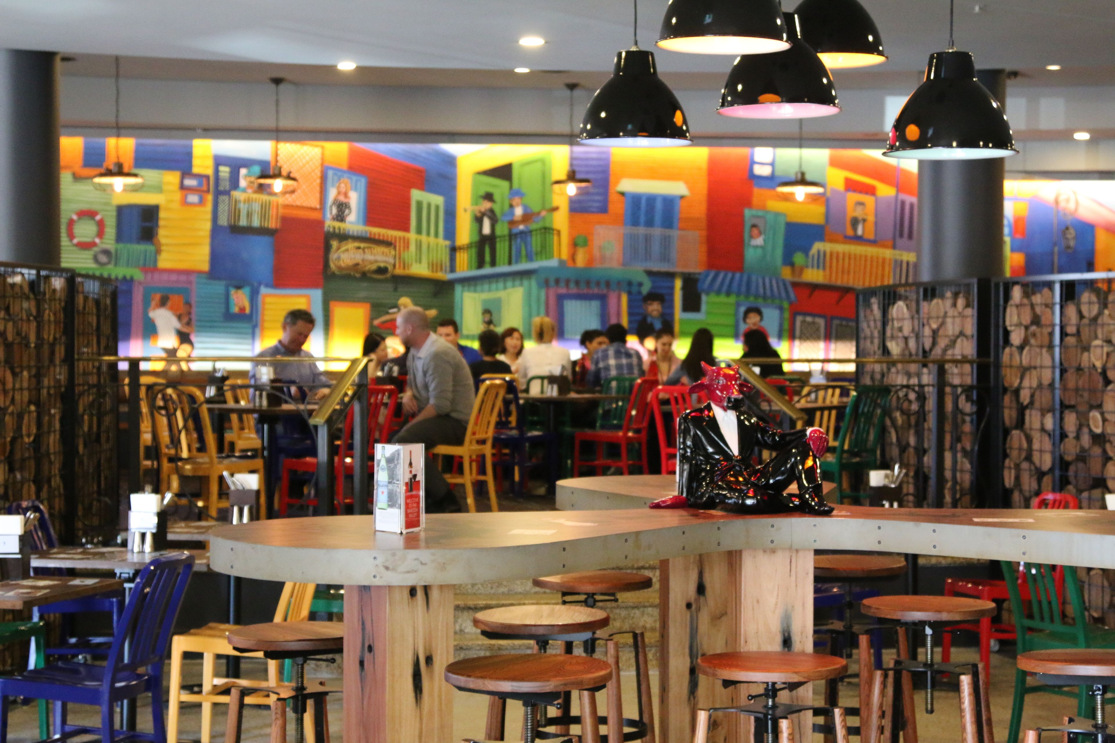 Un restaurante con murales coloridos en las paredes, taburetes de madera en la barra y lámparas colgantes.