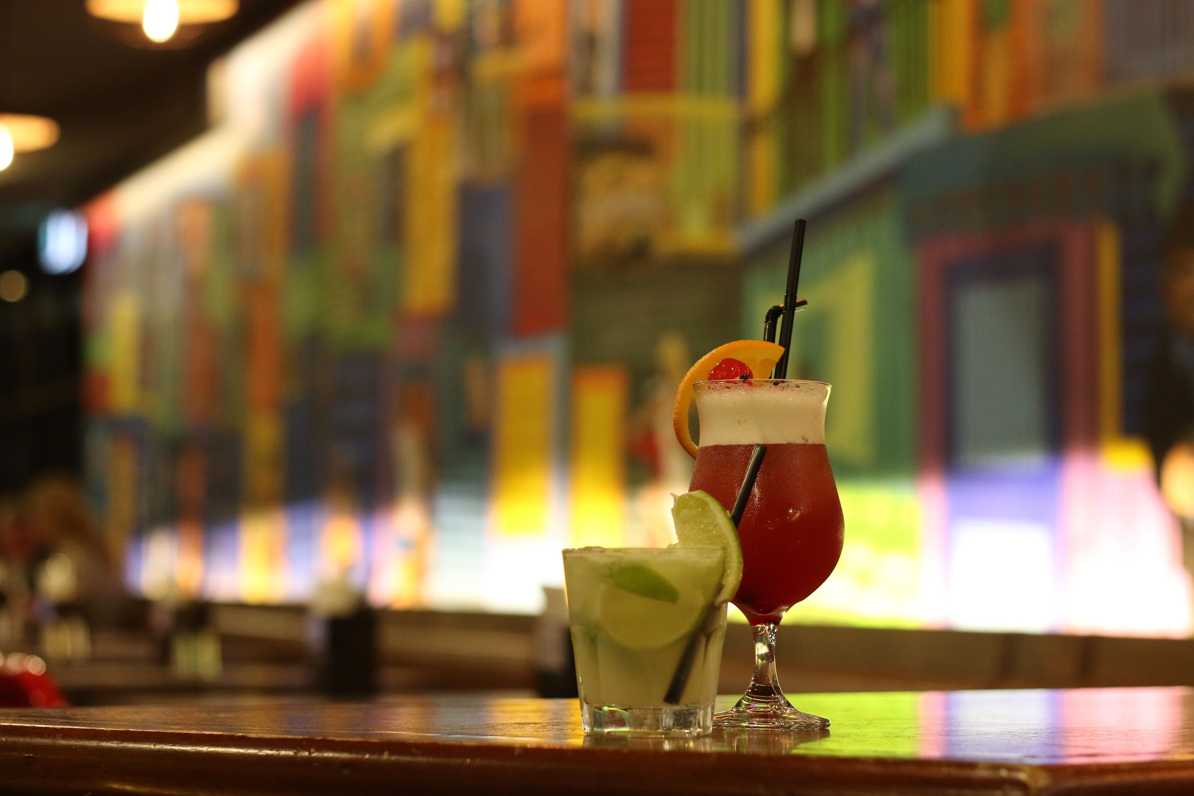 Two cocktails on a bar counter with a colorful mural in the background.