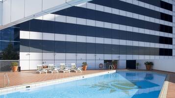 Una piscina con sillas de descanso y plantas en macetas, rodeada por una terraza con baldosas y un edificio moderno con amplias ventanas.
