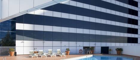 A swimming pool with lounge chairs and potted plants, surrounded by a tiled deck and a modern building with large windows.