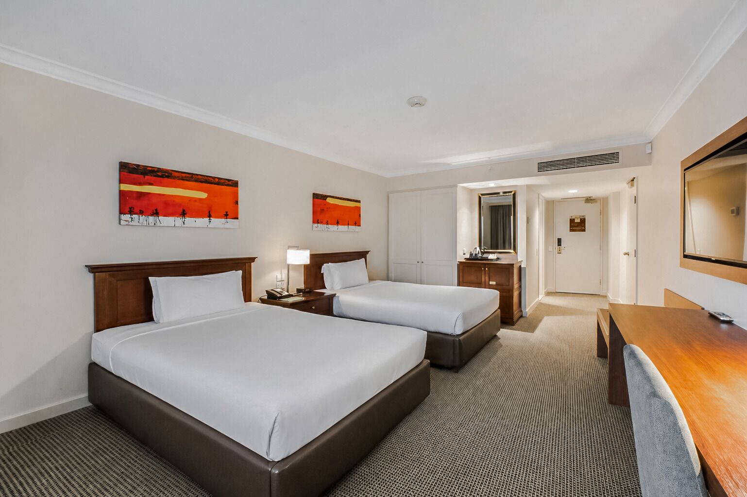 A hotel room with two beds, a desk, a mirror, and artwork on the walls.