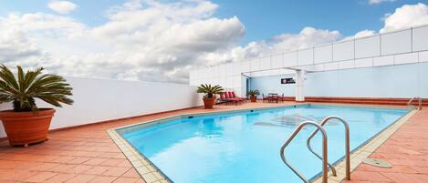 A rooftop swimming pool with a clear blue sky, potted plants, and red chairs.