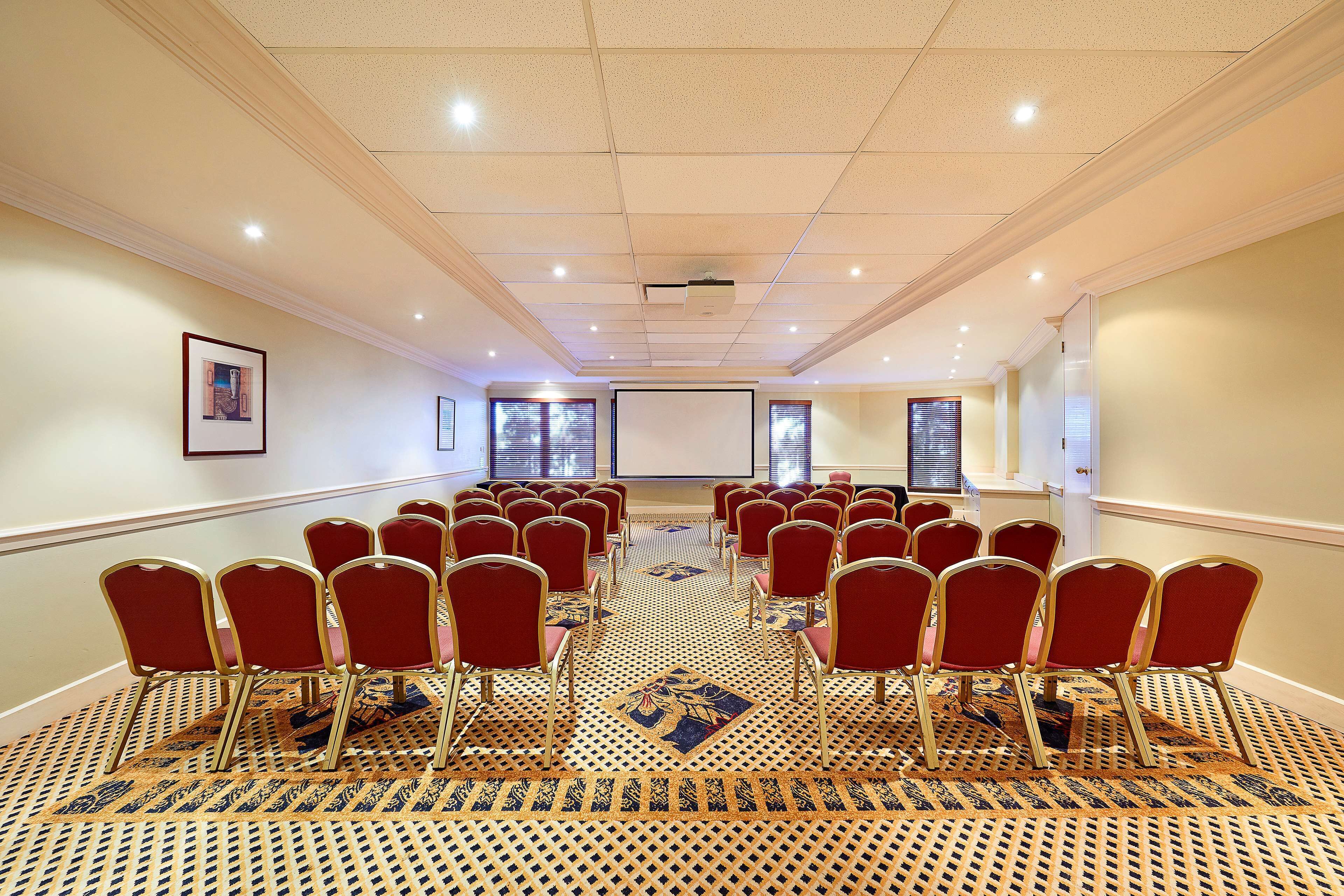 Un salón de conferencias con hileras de sillas rojas, alfombra estampada y una pantalla de proyector.