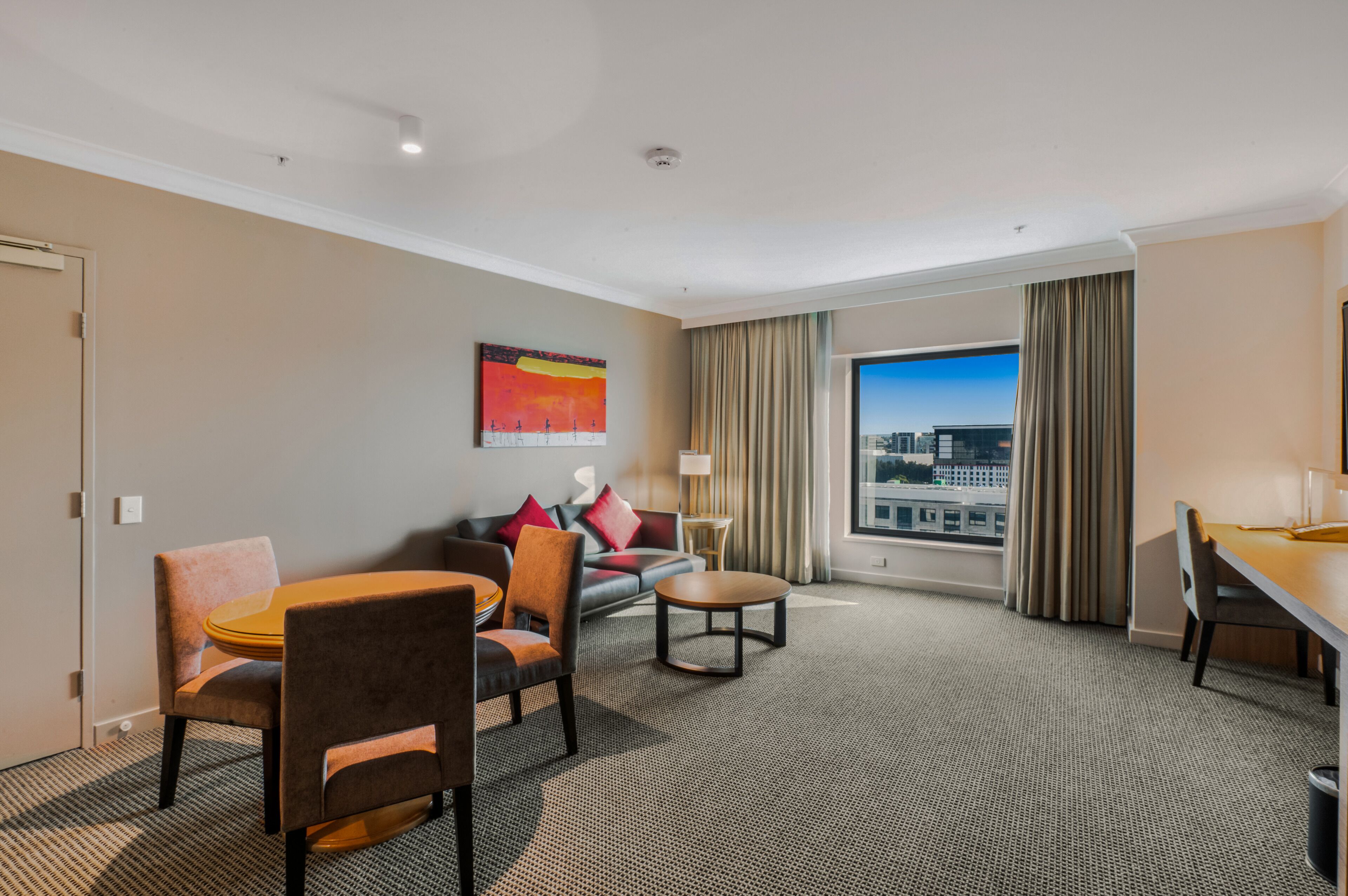 A hotel room with a dining area, a sofa, a coffee table, a lamp, and a large window with a city view.