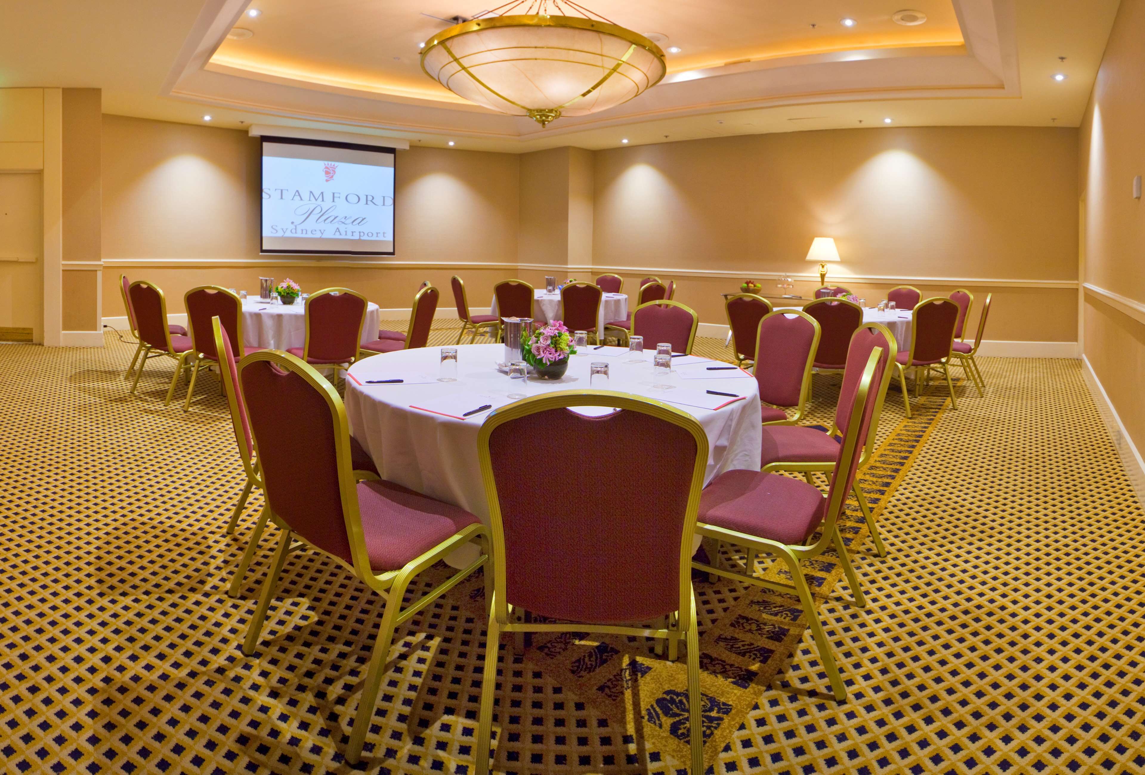 Un salón de conferencias con mesas redondas, sillas y una pantalla de proyector mostrando 'STAMFORI Sydney Airport'.