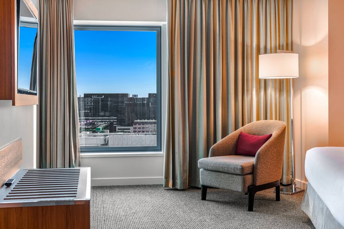 A hotel room with a bed, a chair, a desk with a lamp, a window with curtains, and a view of the cityscape.