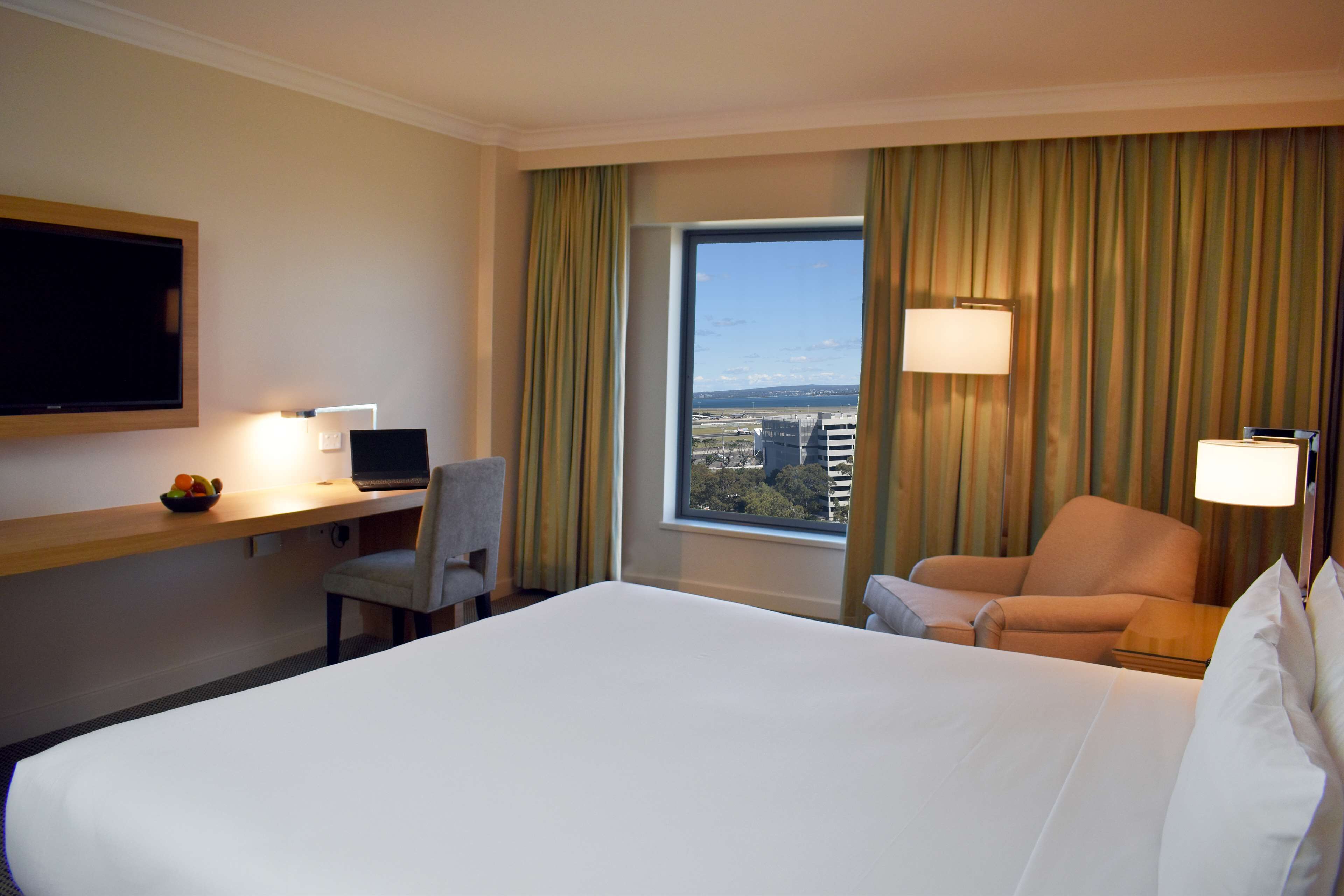 A hotel room with a large bed, a desk with a computer, a chair, a TV mounted on the wall, and a view of the city through a large window.
