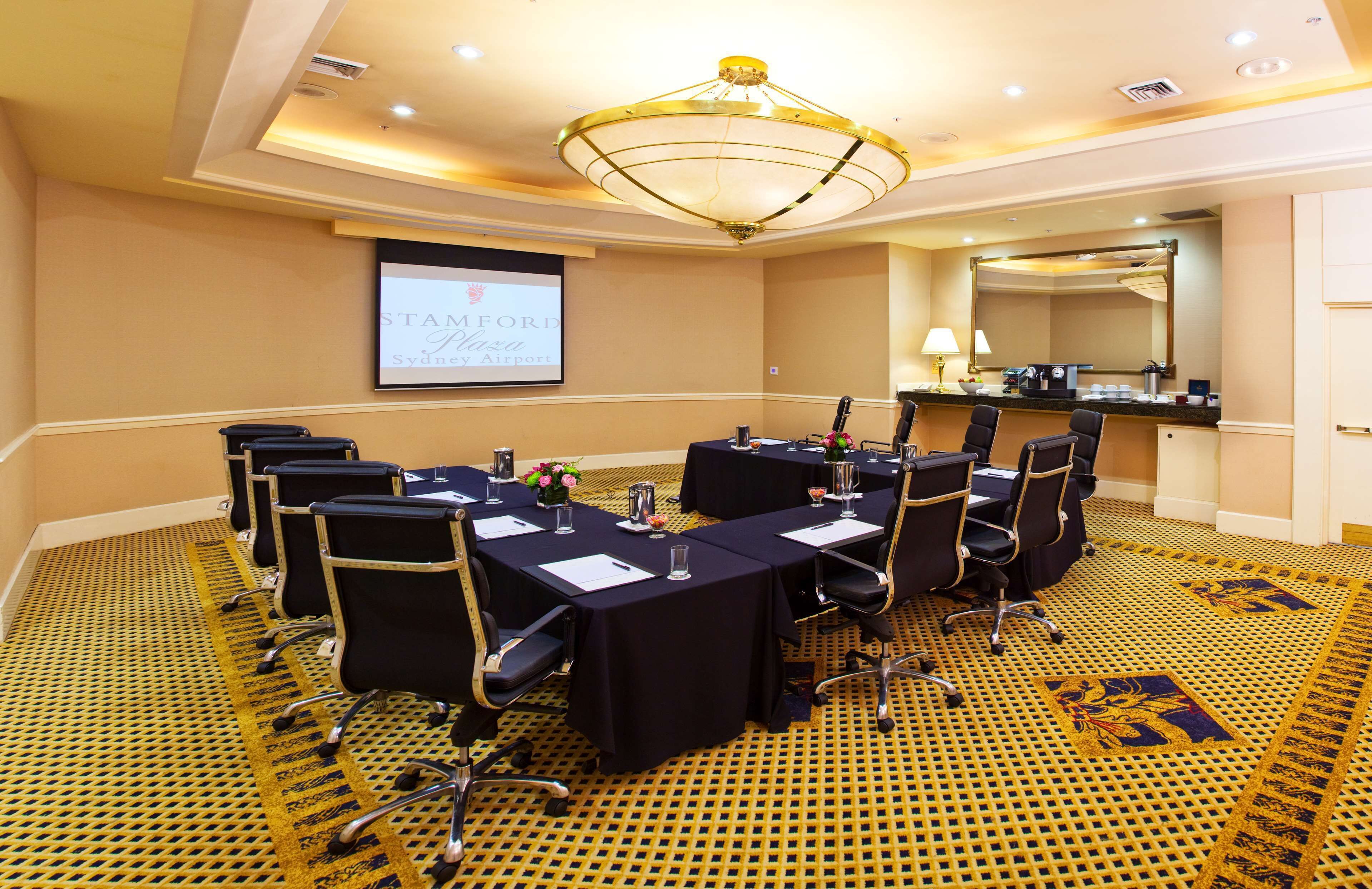 A conference room with a projector screen displaying 'STAMFORD Sydney,' a long table with chairs, a carpeted floor, and a chandelier.