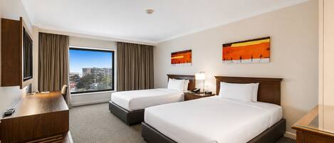 Habitación de hotel con dos camas, un escritorio, un televisor y un ventanal con vista a la ciudad.