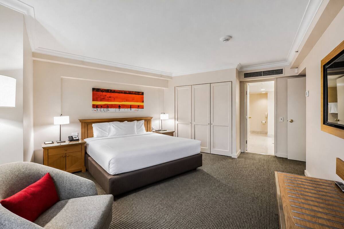 A hotel room with a bed, a desk, a chair, a painting, and a wardrobe.