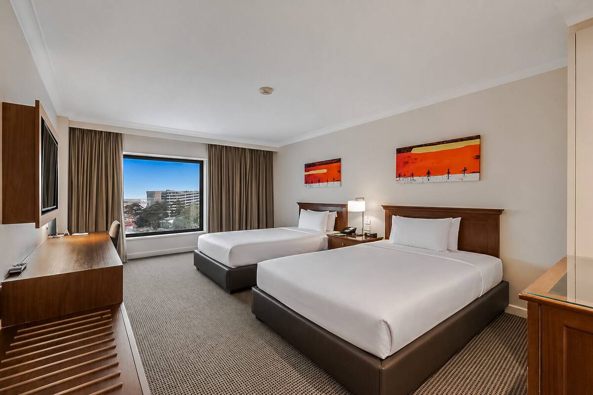 A hotel room with two beds, a desk, a TV, and a large window with a city view.