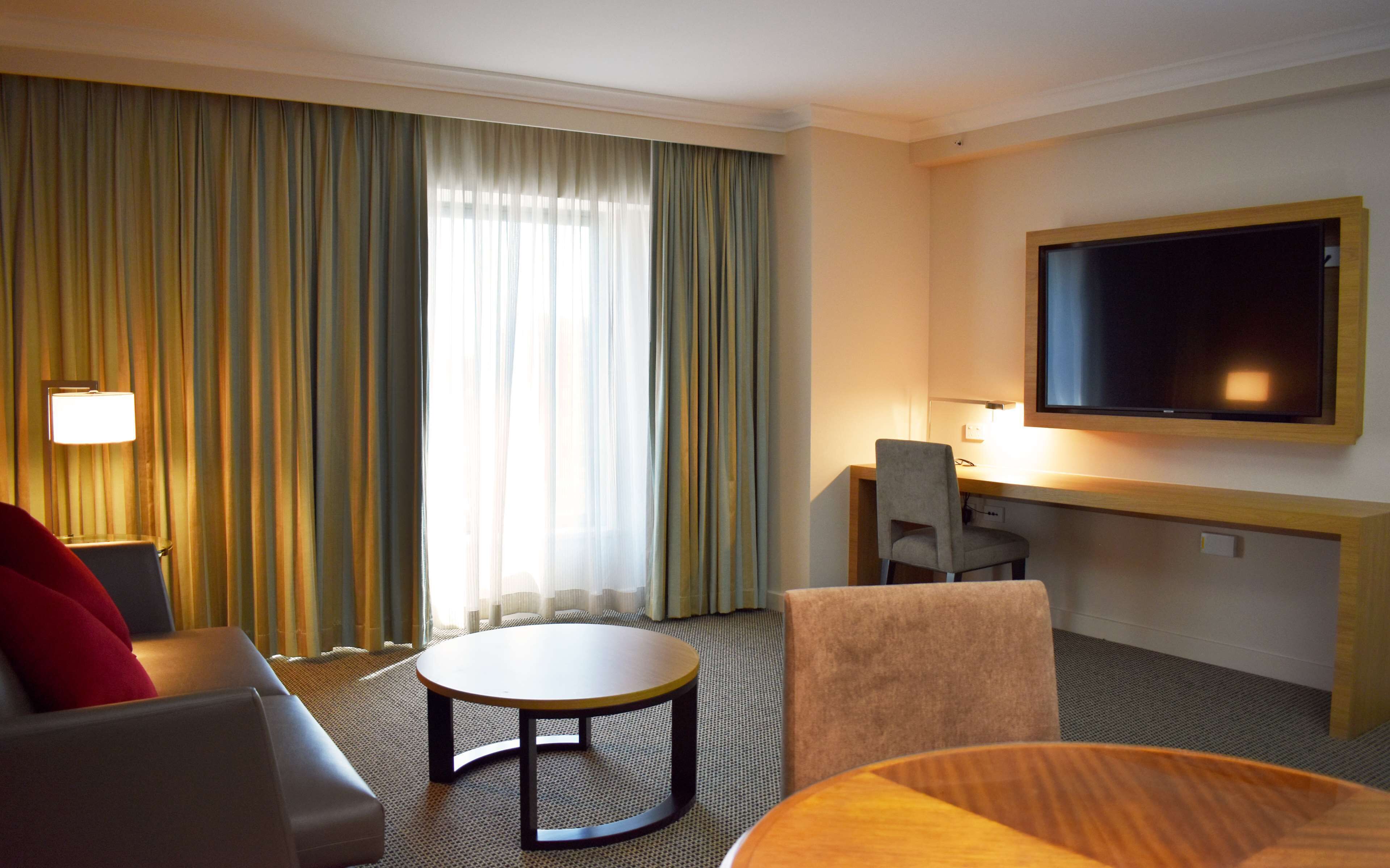 A hotel room with a flat-screen TV, a desk, a chair, a sofa, and a round table.