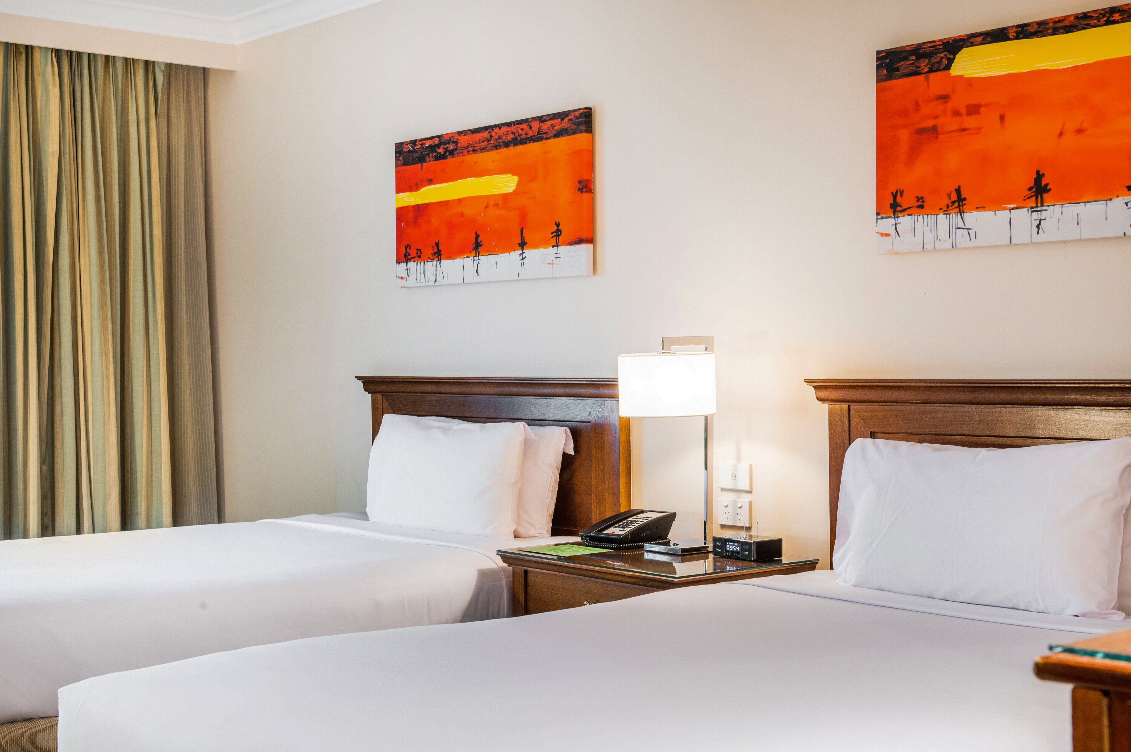 Habitación de hotel con dos camas, una mesita de noche con lámpara y dos cuadros abstractos en la pared.