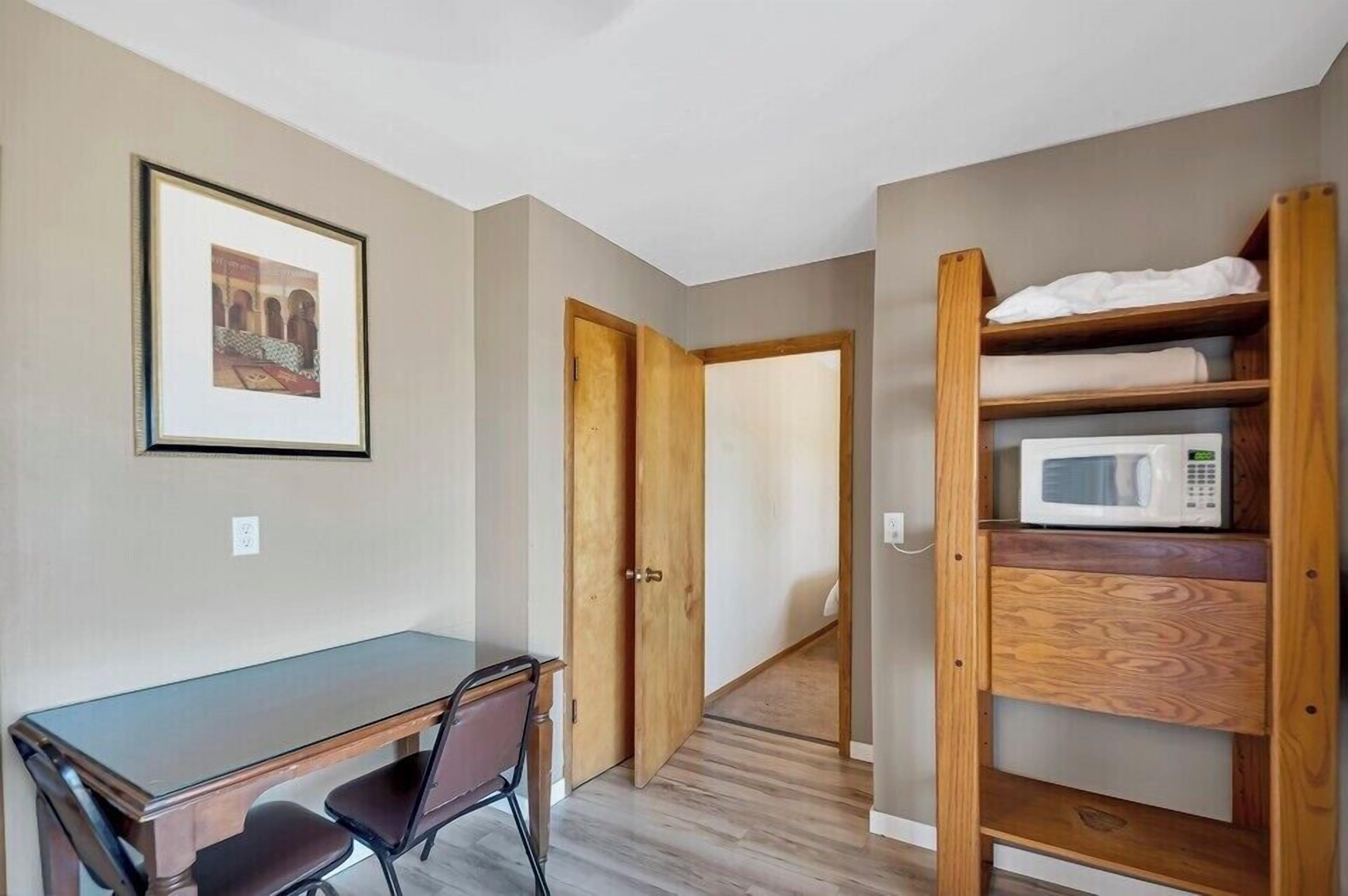 A room with a wooden desk, a chair, a framed picture on the wall, a microwave on a wooden shelf, and a door leading to another room.
