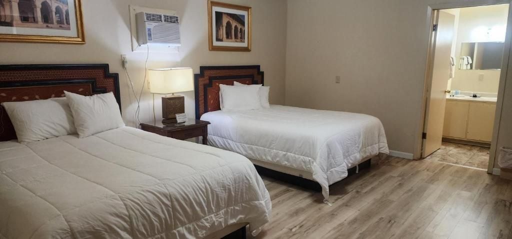 Habitación de hotel con dos camas, una mesita de noche con lámpara, aire acondicionado instalado en la pared y una puerta que da a un baño.