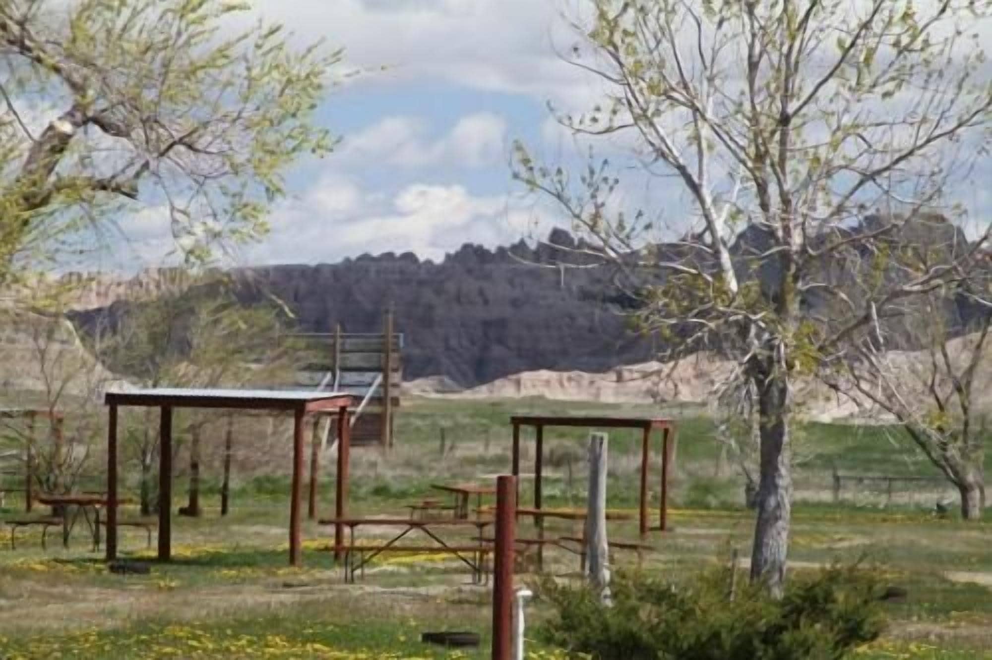 Un cadre extérieur rustique, avec des tables de pique-nique et une structure en bois, entouré d’arbres et dominé par un paysage montagneux.