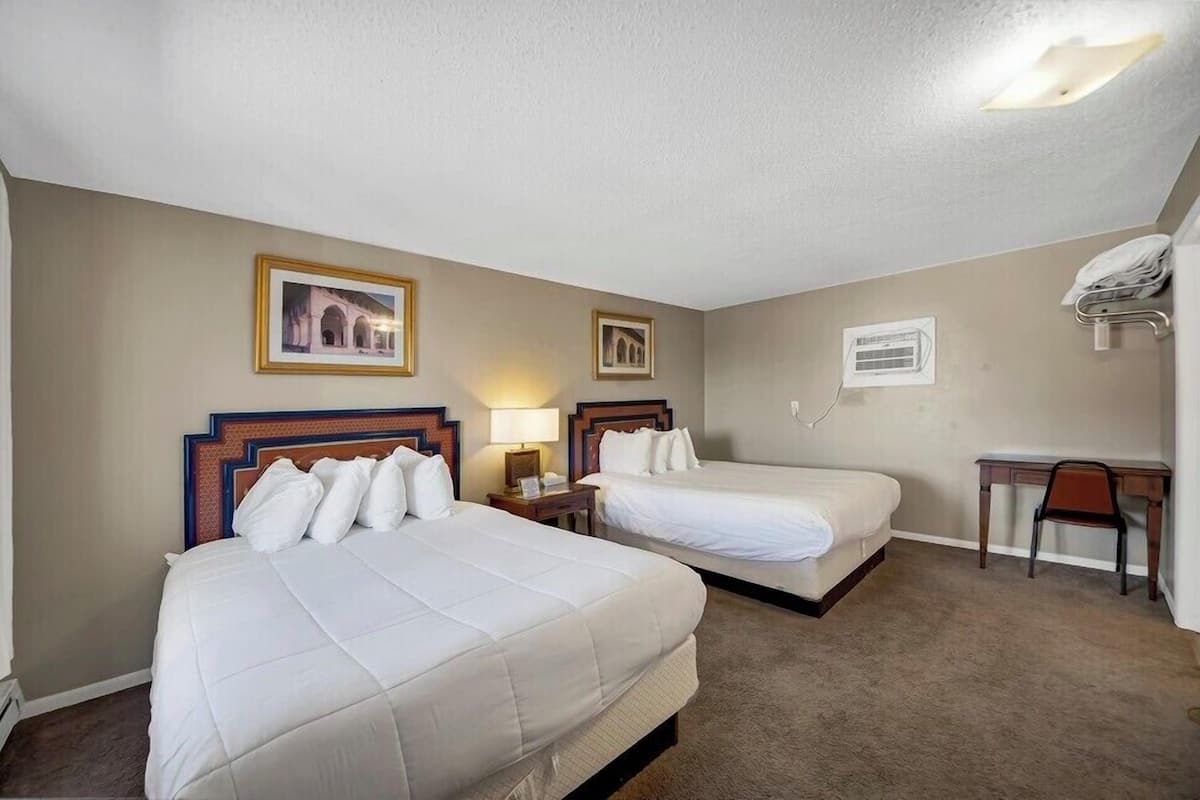 Quarto de hotel com duas camas, uma mesa, uma cadeira e uma luminária.