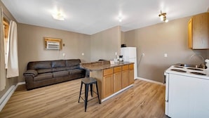 Un espace de vie compact comprenant un coin cuisine, un canapé en cuir marron, un petit tabouret en bois et une cuisinière blanche.