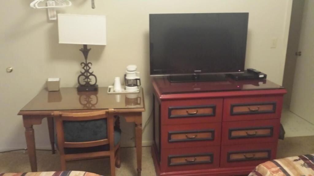 A hotel room with a wooden desk, a television on a stand, a red dresser with drawers, a lamp, and a coffee maker.