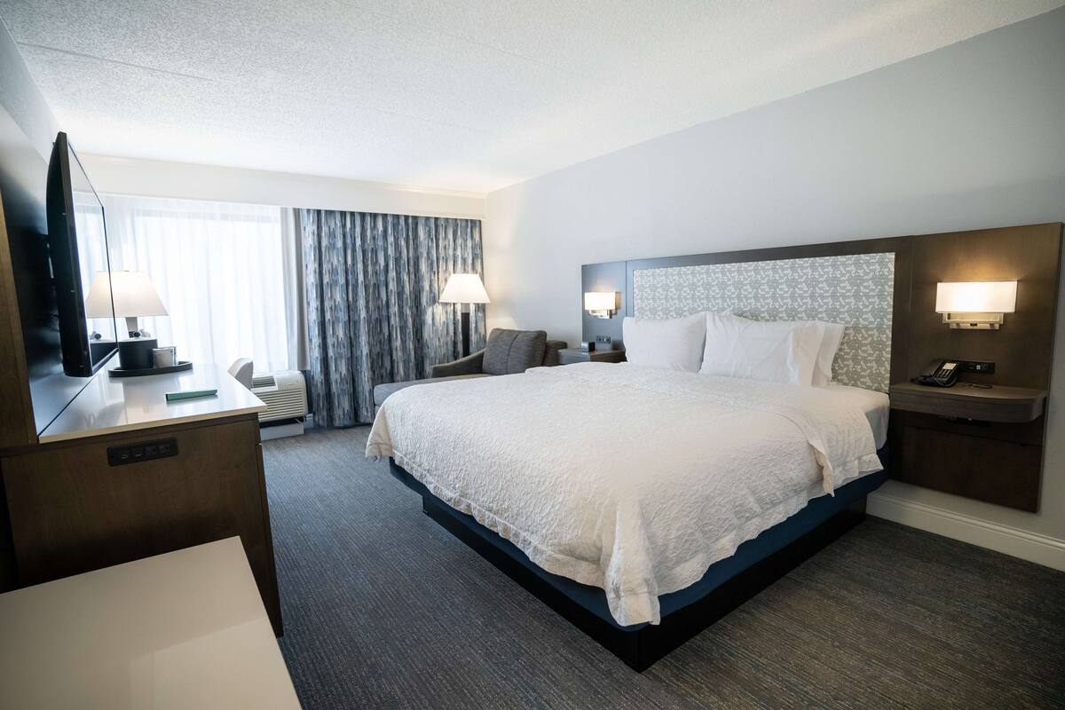 Quarto de hotel com uma cama grande, abajures, uma escrivaninha com televisão de tela plana e uma janela ampla com cortinas.