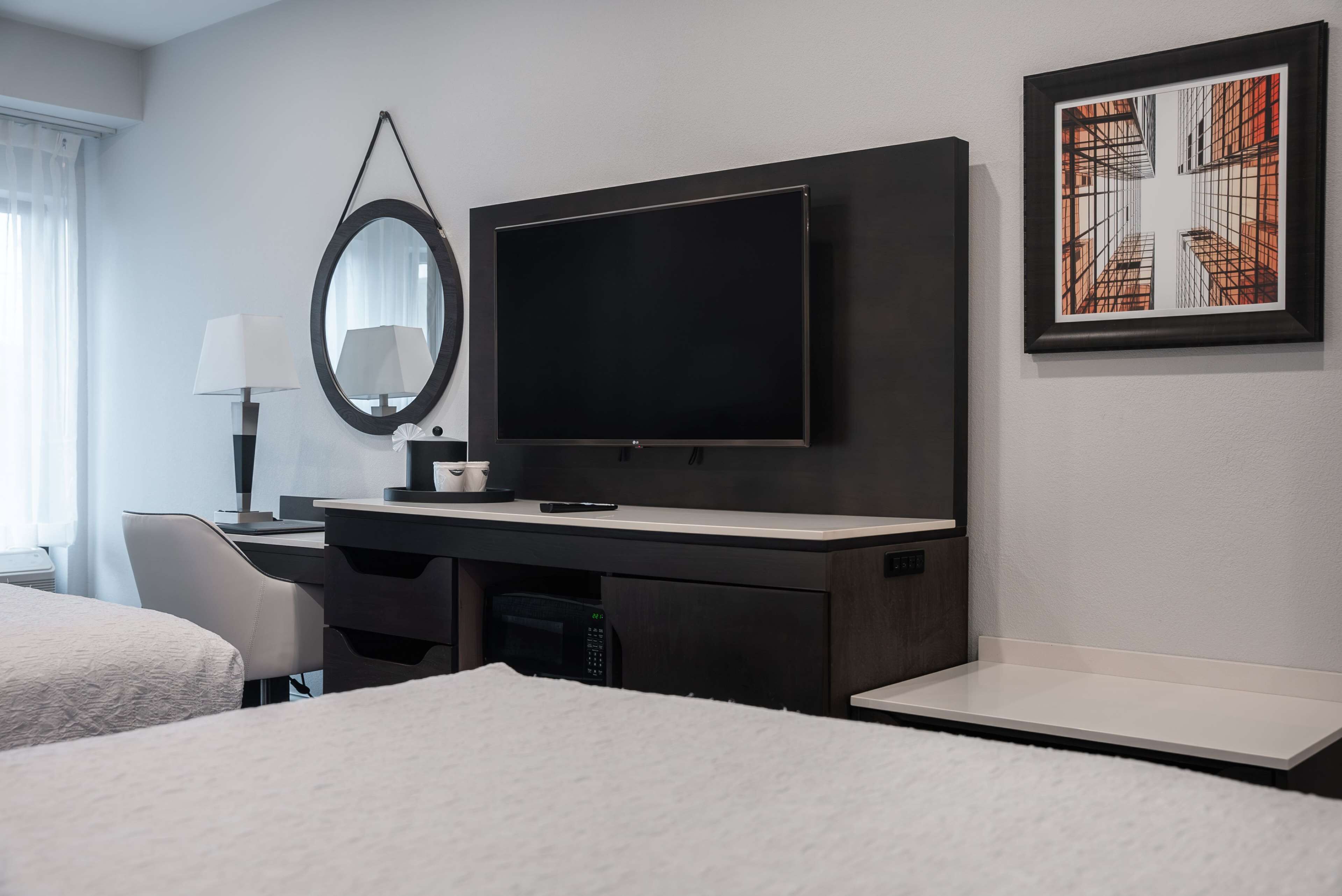 A modern hotel room with a flat-screen TV, a desk with a lamp, a large mirror, and a framed artwork on the wall.