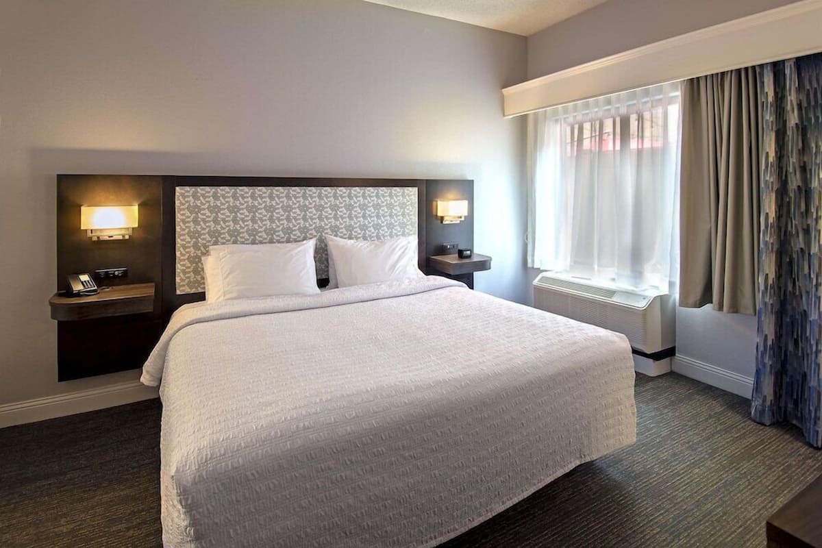 Quarto de hotel com uma cama grande, duas mesas de cabeceira com abajures, uma janela com cortinas e um ar condicionado fixado na parede.