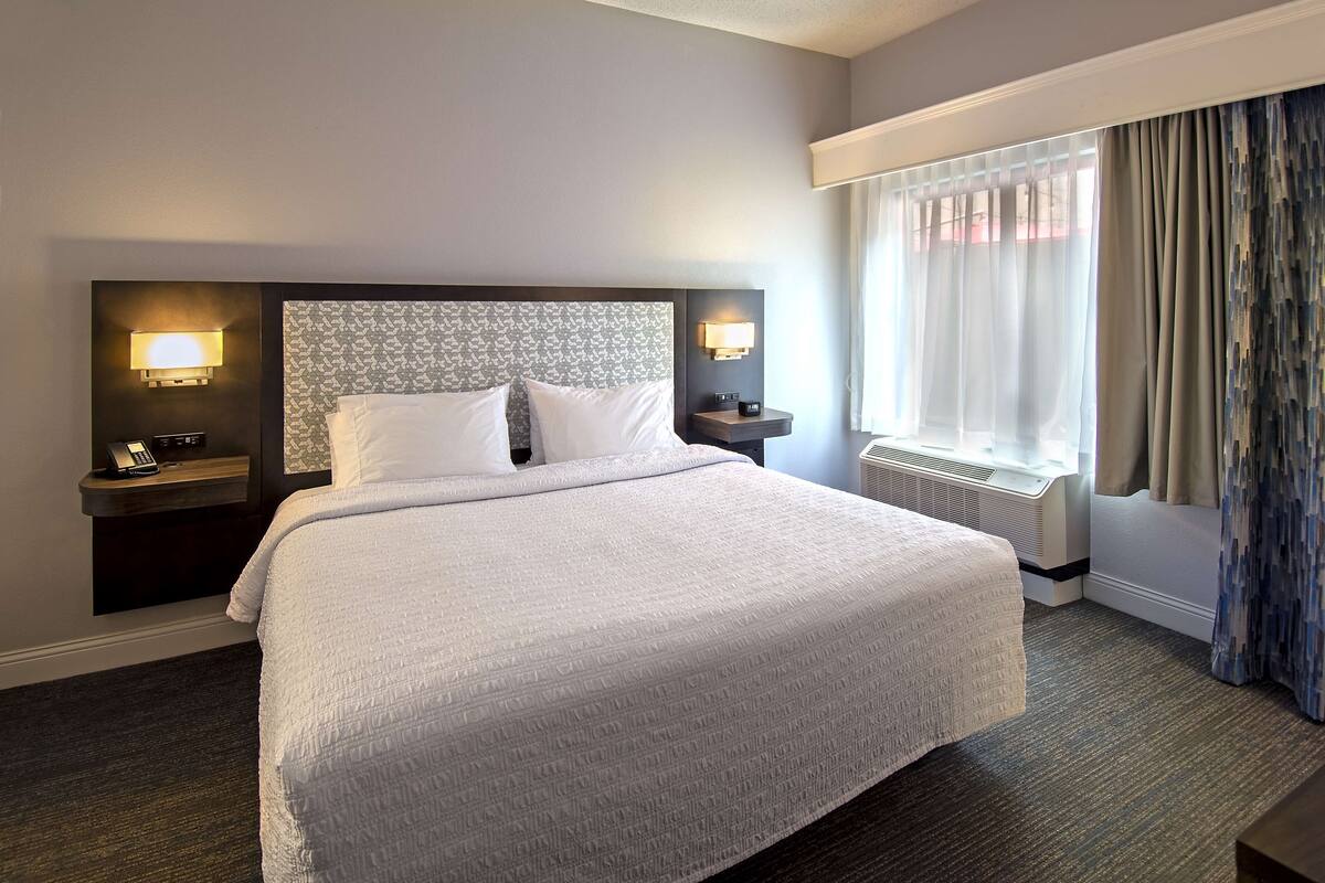 Quarto de hotel com uma cama grande, duas mesas de cabeceira com luminárias, uma janela com cortinas e um radiador.
