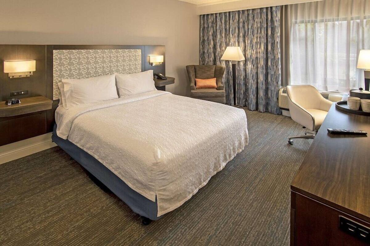 Quarto de hotel com uma cama grande, uma escrivaninha, uma cadeira, uma mesa de cabeceira e uma luminária.