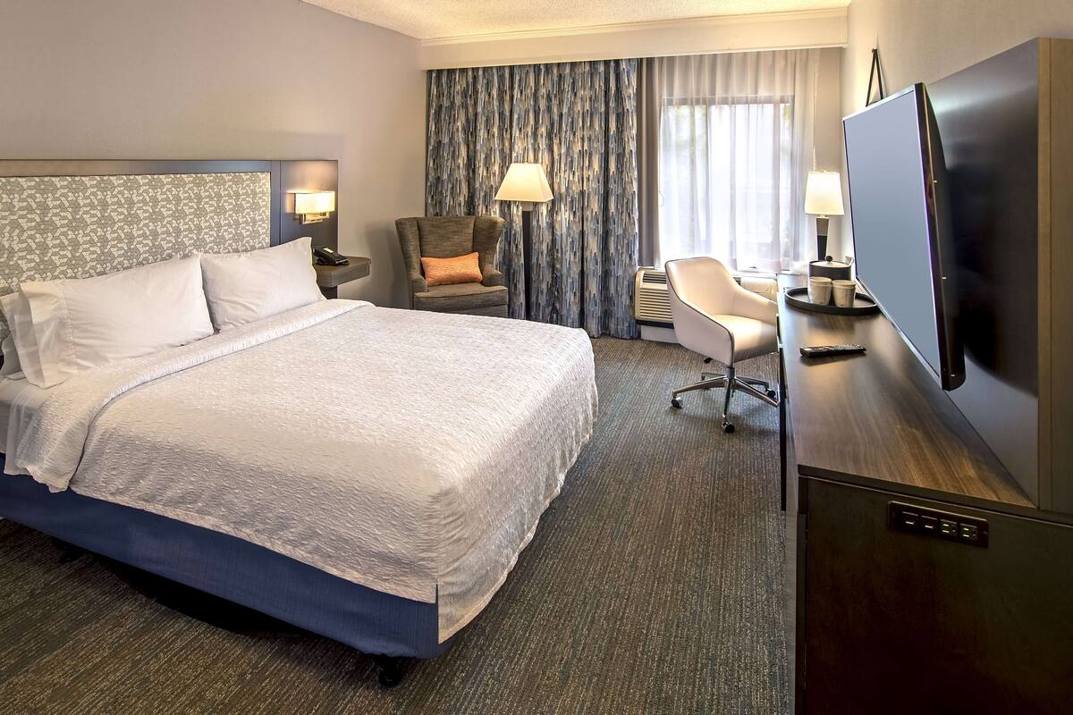 Quarto de hotel com uma cama grande, uma escrivaninha com televisão de tela plana, uma cadeira e um abajur.