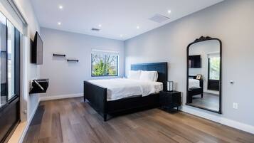 A modern bedroom with a large bed, a TV mounted on the wall, a large mirror, and a window with a view of trees.