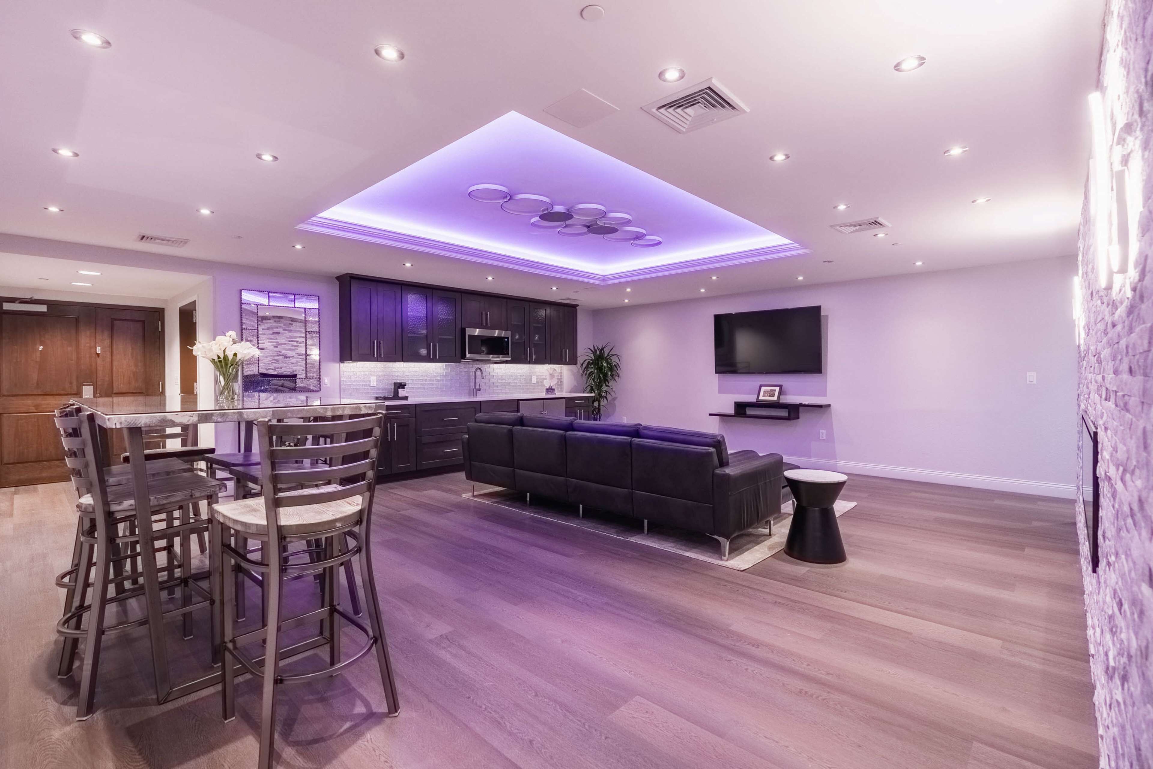 A modern living room with a dining area, a kitchen, and a flat-screen TV mounted on the wall.