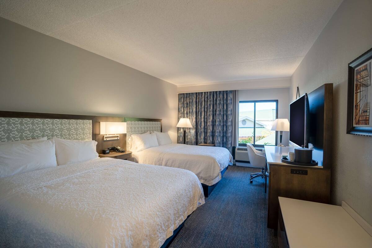 Quarto de hotel com duas camas, uma escrivaninha com computador, televisão e janela com cortinas.