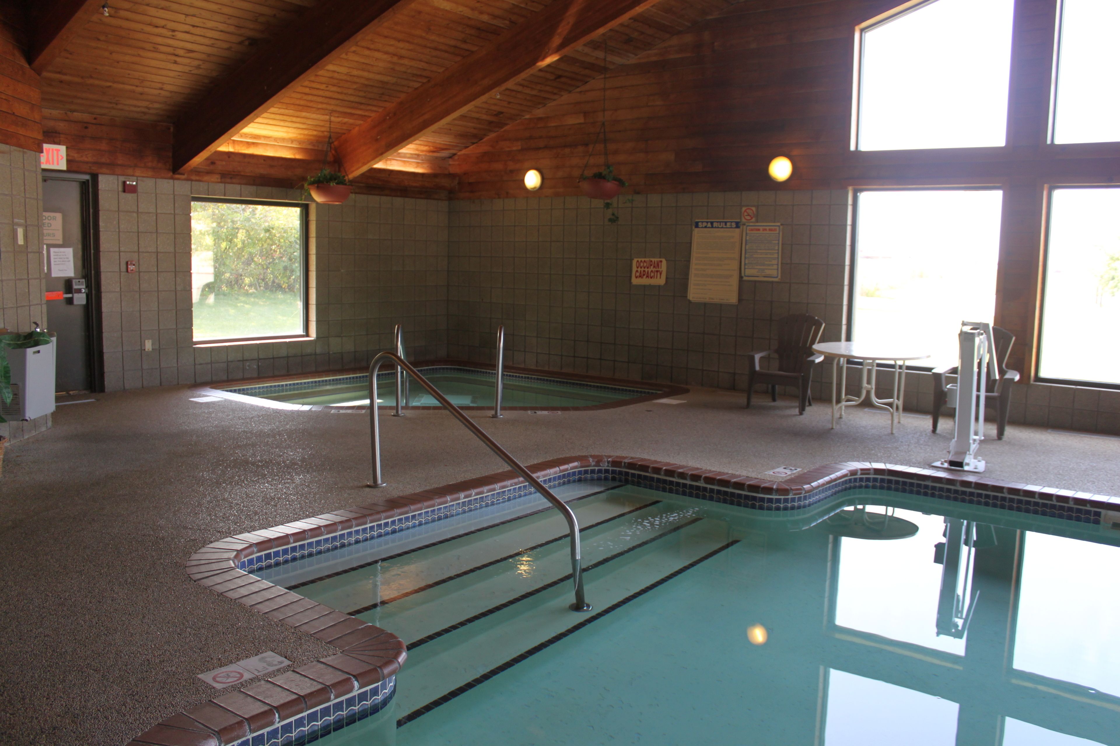 An indoor swimming pool with a tiled floor, a metal ladder, and a seating area with chairs and a table.