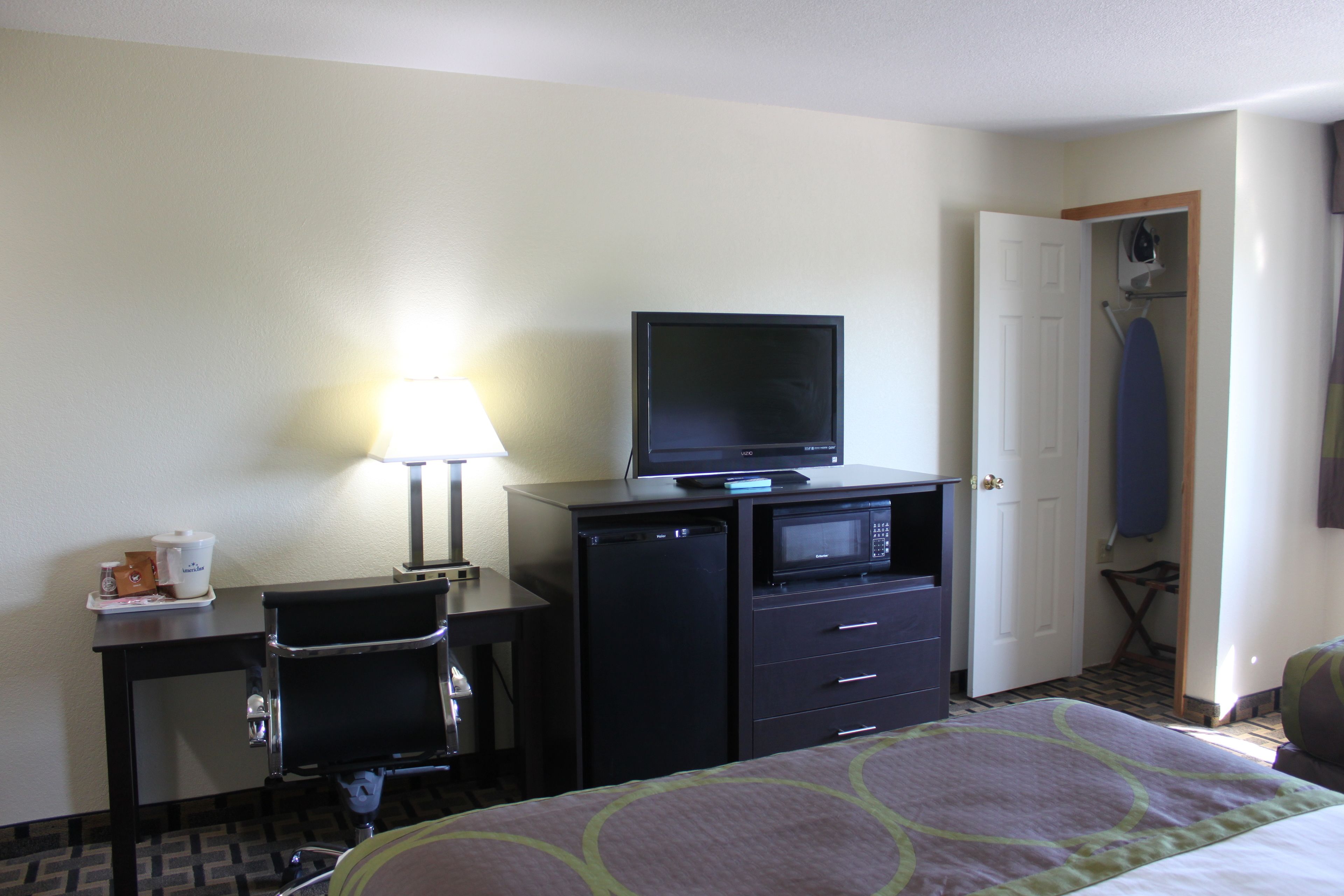 A hotel room with a bed, a desk with a computer and a television, a small refrigerator, and a lamp.