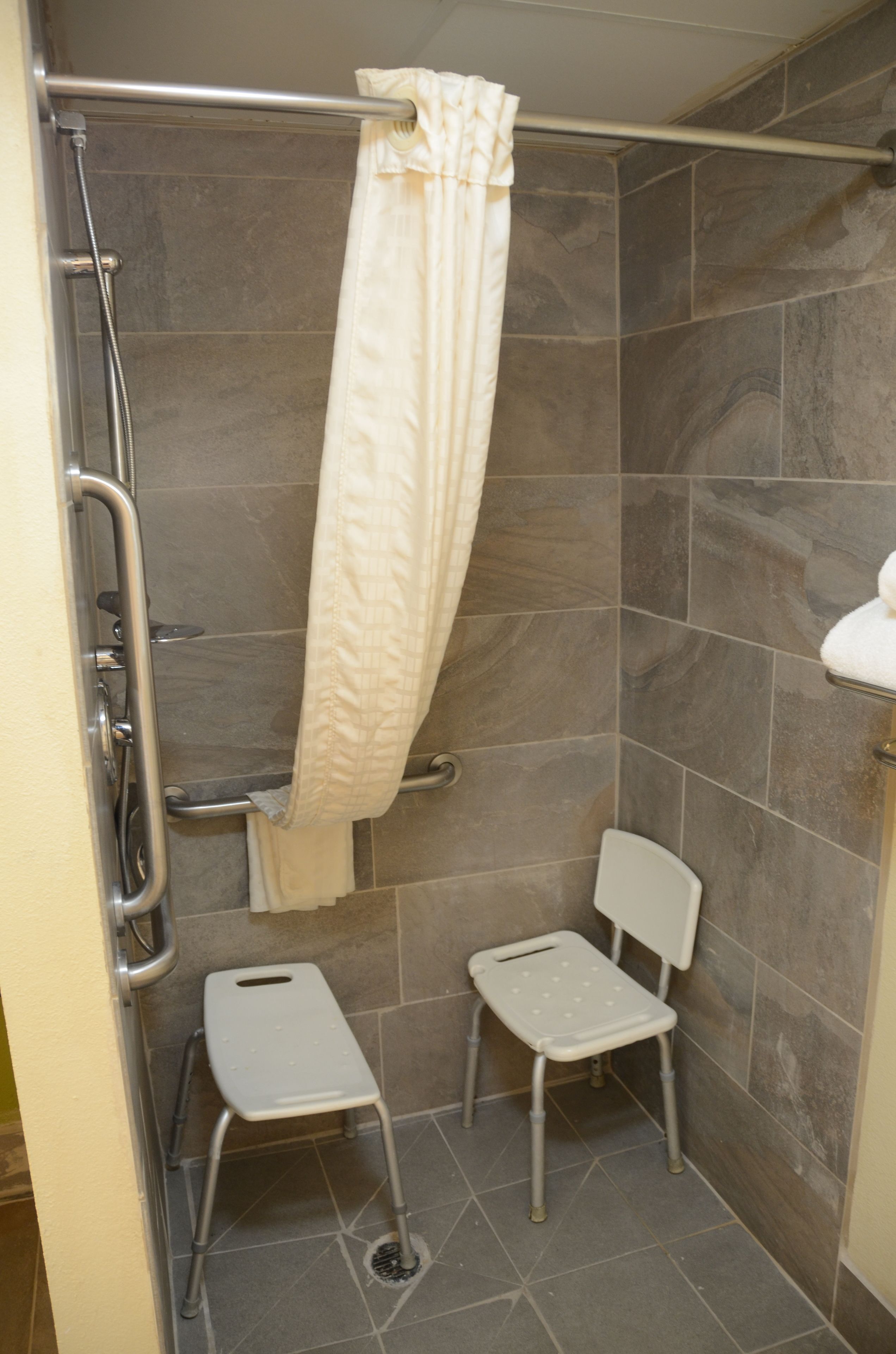 Un espace douche équipé d’une barre d’appui, d’un rideau de douche blanc et d’un petit tabouret blanc.