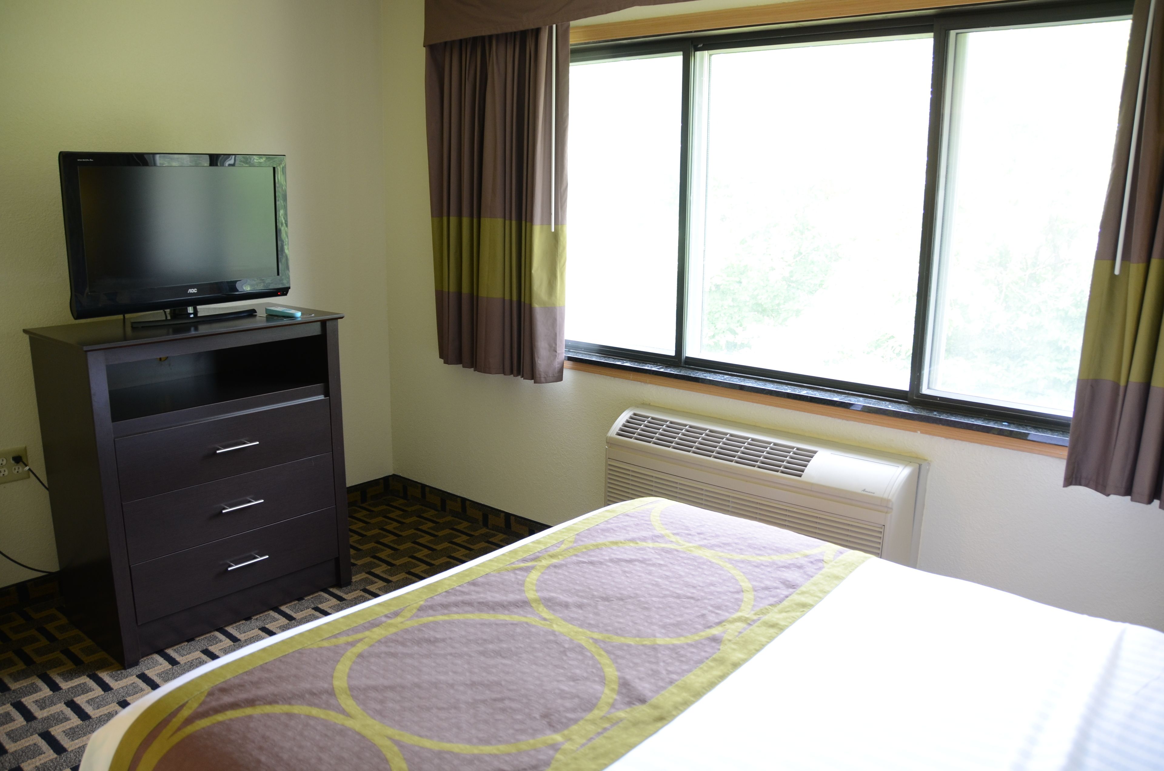 Habitación de hotel con cama, televisor en una cómoda, ventana con cortinas y radiador.
