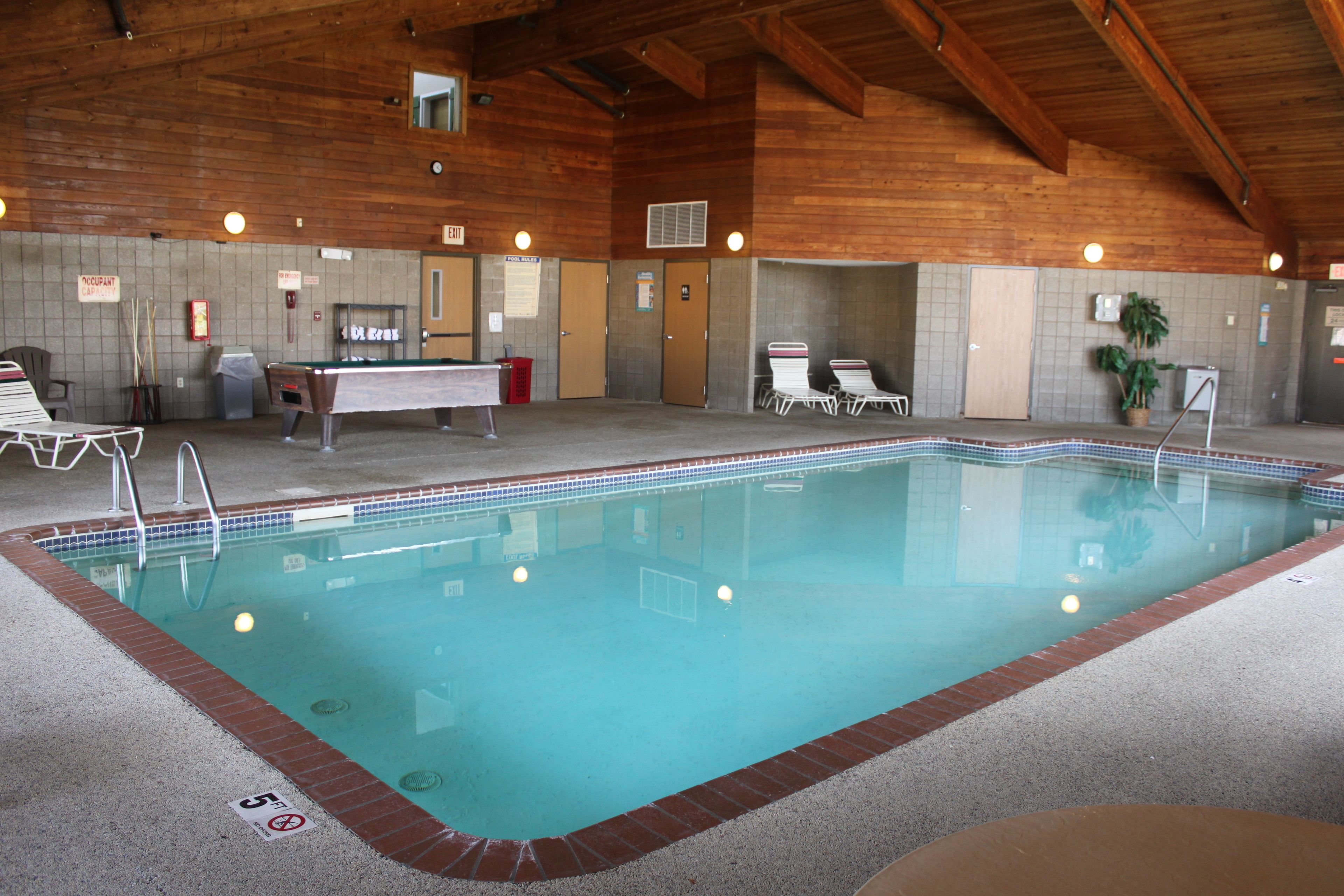An indoor swimming pool with a wooden ceiling, a pool table, and lounge chairs.