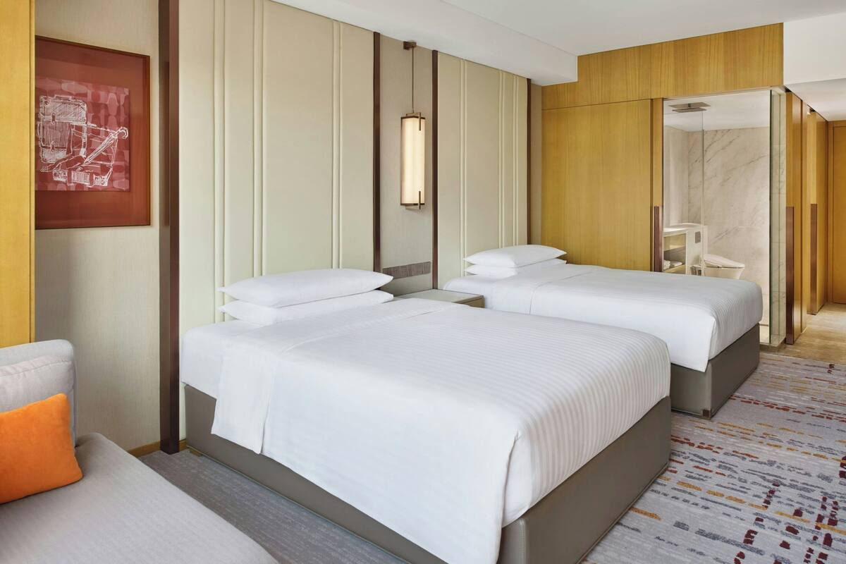 A hotel room with two beds, a sofa, a framed picture, a pendant light, and a bathroom with a shower and toilet.