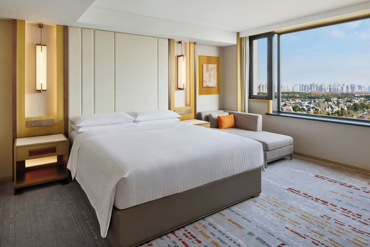 A hotel room with a large bed, a sofa, a nightstand, a wall-mounted lamp, and a large window with a city view.