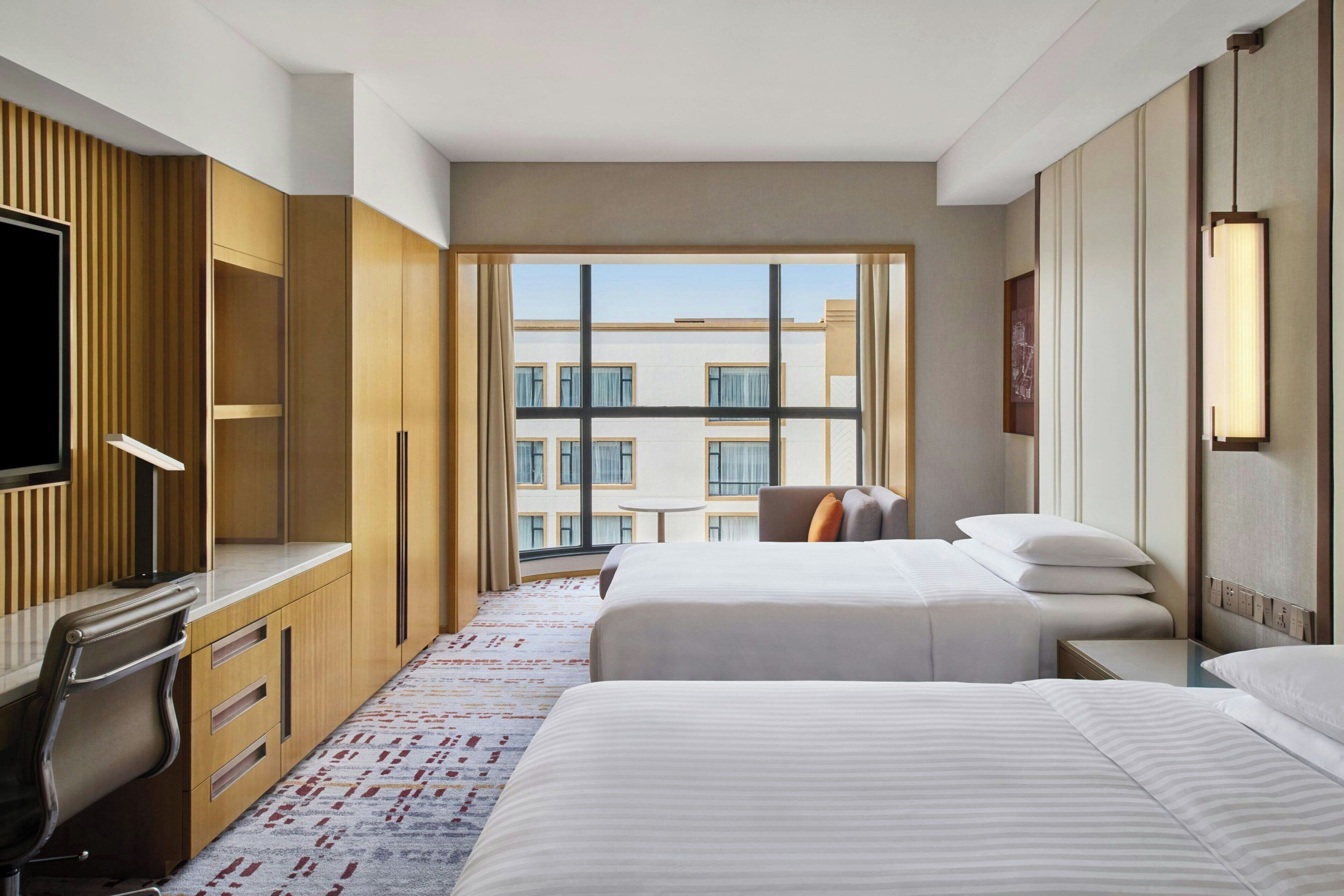 A hotel room with a large window, a desk, two beds, and a TV.