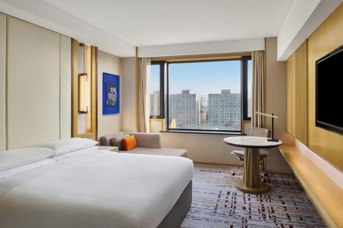 A hotel room with a large bed, a sofa, a small table, a TV, and a window with a city view.