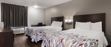 A hotel room with two beds, a desk, a chair, a radiator, and a window with curtains.