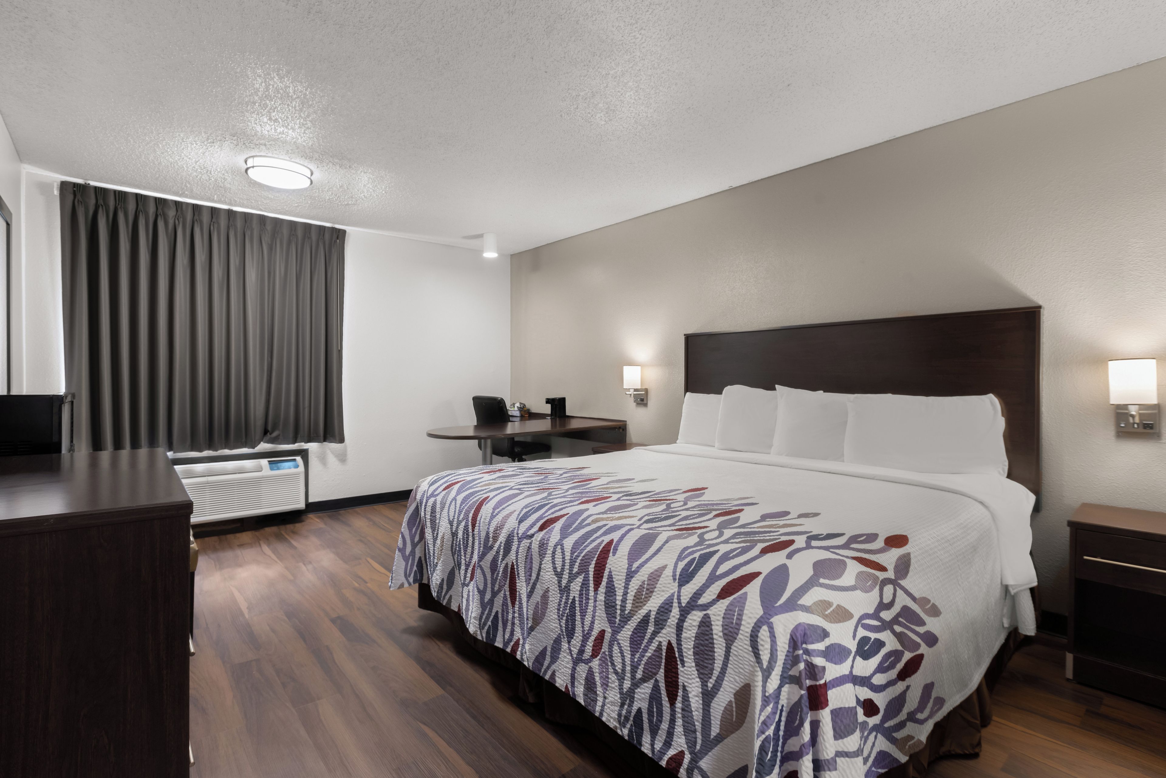 Una camera d'albergo con un letto, una scrivania, una sedia, una televisione, un termosifone e una finestra con tende.
