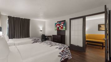 Quarto de hotel com duas camas, uma escrivaninha, uma cadeira, televisão e um sofá.