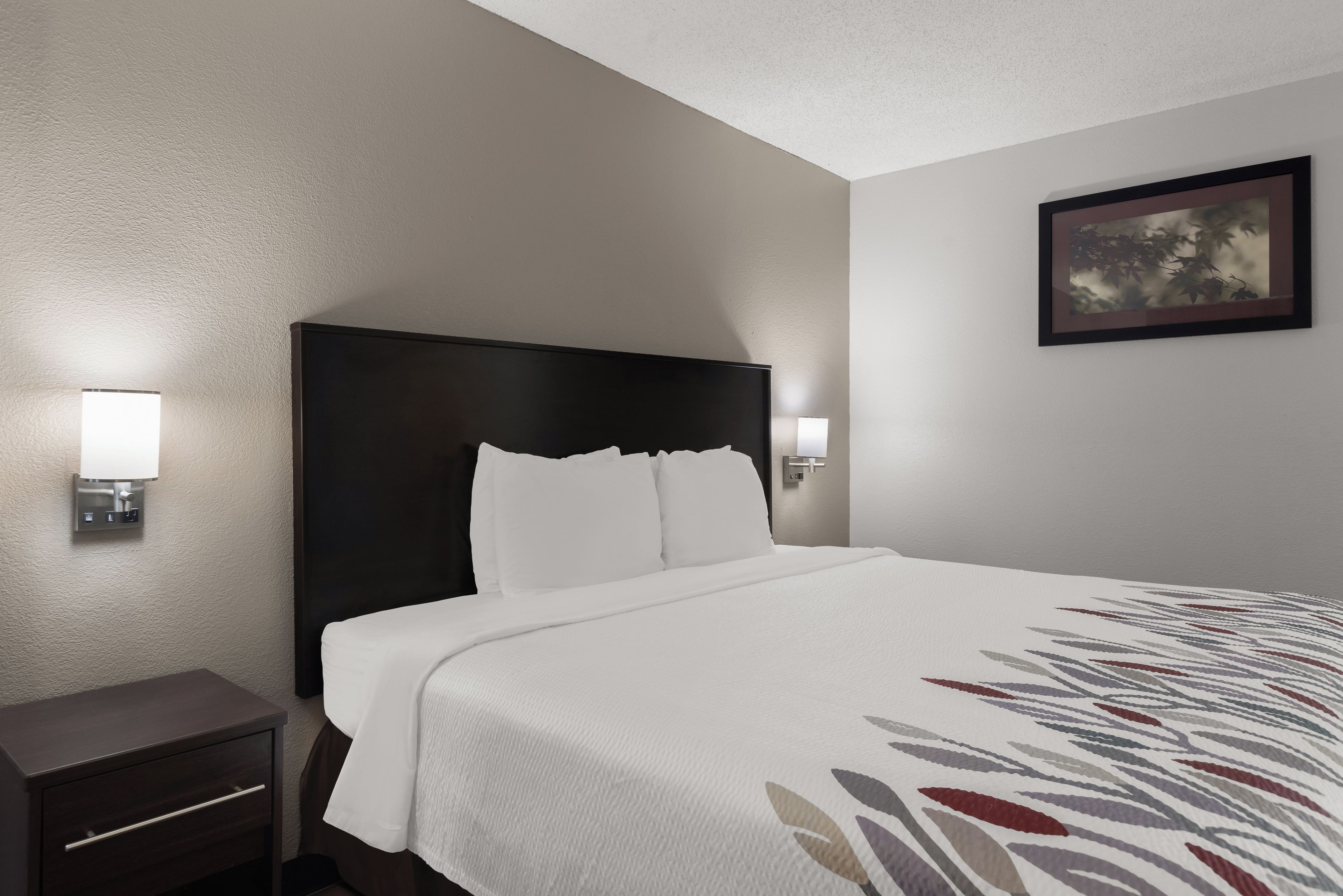 A hotel room with a bed, nightstand, wall-mounted lights, and a framed picture.