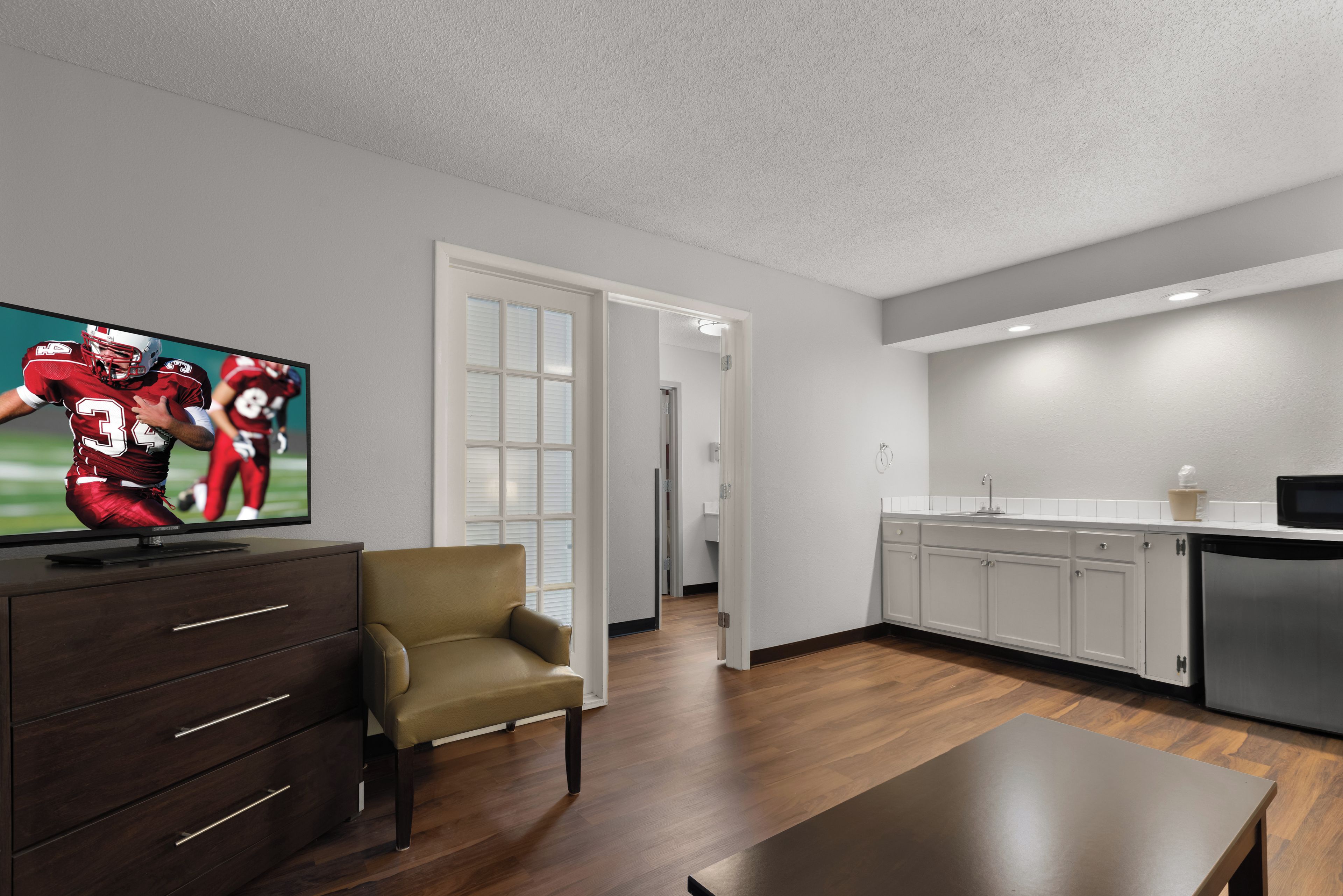 Una sala moderna con un televisor de pantalla plana, un mueble de madera oscura, un sillón beige y una cocina con armarios blancos y un refrigerador de acero inoxidable.